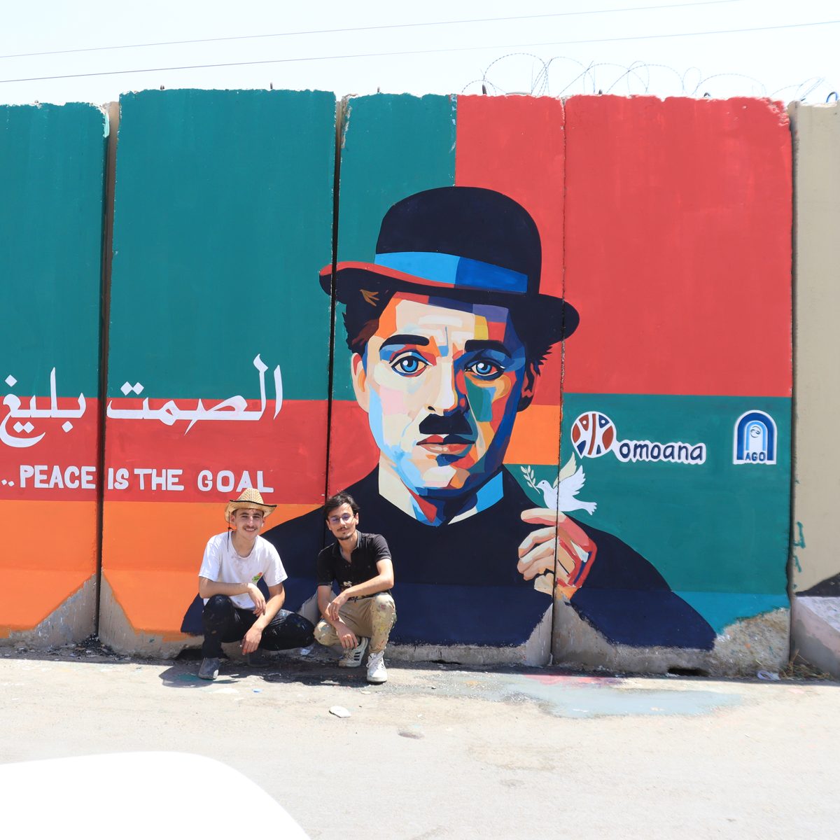 Two young artists in front of their “Silence is eloquent… peace is the goal” mural with the Omoana and AGO logos
